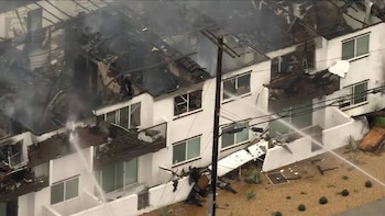 Edificio de apartamentos de varios pisos con techo colapsado y daños por incendio, mientras mangueras de bomberos rocían agua sobre la estructura