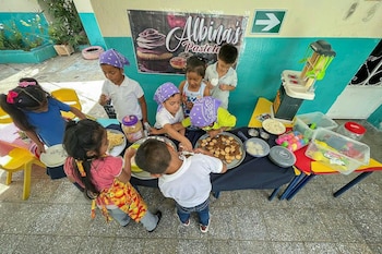 Varios niños reunidos alrededor de una mesa con galletas, platillos de juguete y una cocina de plástico bajo un cartel de repostería