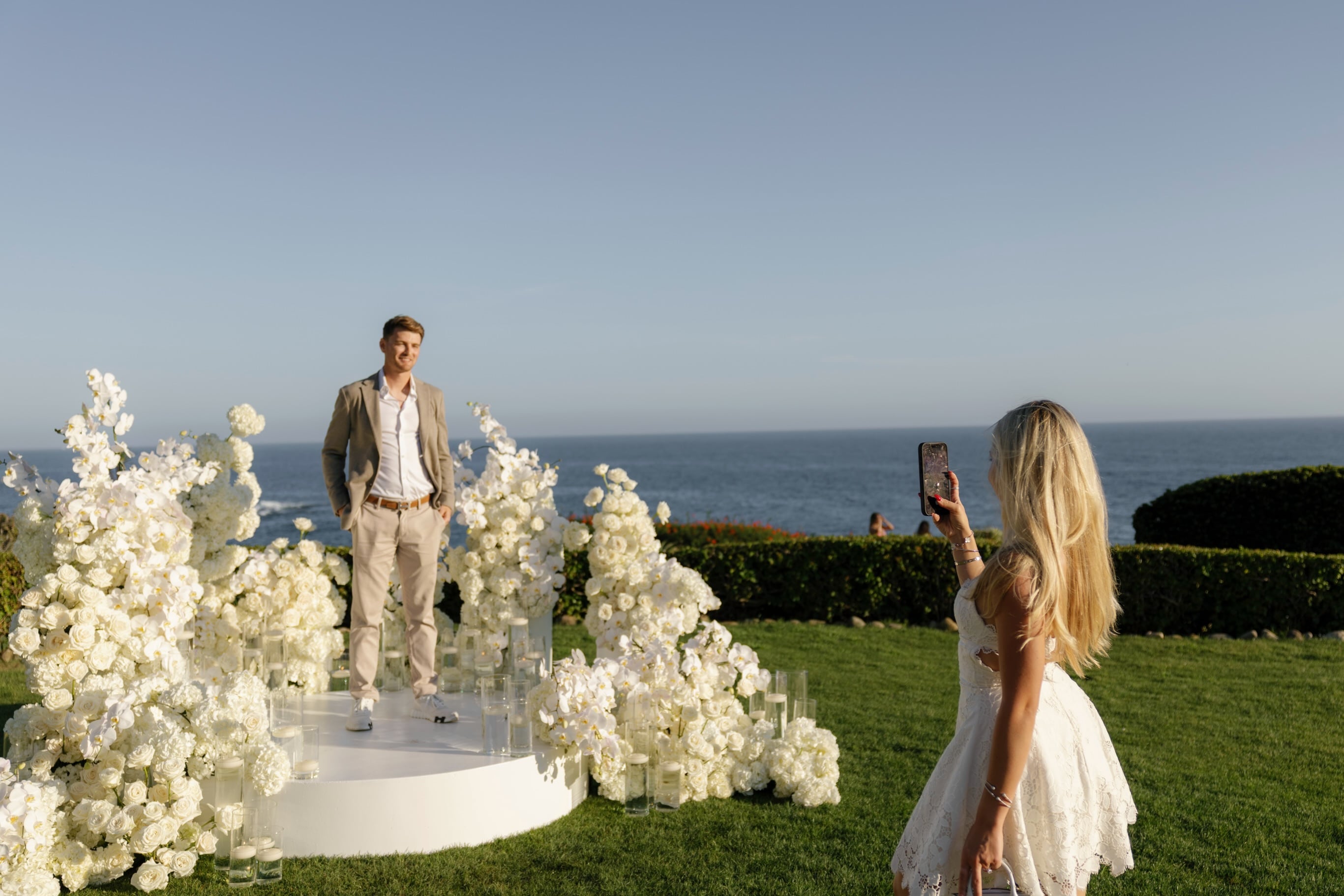 El expiloto de F1 Logan Sargeant y su pareja celebran su compromiso en un evento al aire libre decorado con flores blancas y vista al mar.