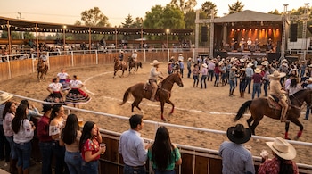 Charros a caballo, mujeres con vestidos tradicionales y asistentes se encuentran en un ruedo de arena junto a un escenario con músicos.