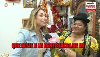 Una mujer rubia con chaqueta azul habla frente a un micrófono rodeada de adorno religioso y otra mujer vestida de amarillo