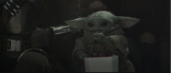 Grogu, la criatura de piel verde y orejas grandes, come un dulce de una caja blanca llena de otros dulces de colores, sentado junto a un brazo con armadura oscura