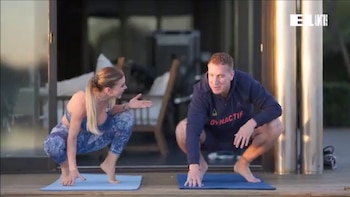 Una mujer y un hombre, ambos descalzos, hacen una sentadilla sobre colchonetas de yoga en una terraza al aire libre