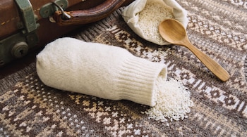 Calcetín blanco con arroz, granos de arroz esparcidos, saco de tela con arroz, cuchara de madera, manta tejida, y una maleta de cuero.