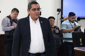 Un hombre con gafas, camisa blanca y saco oscuro camina frente a varias personas que sostienen cámaras y teléfonos en una sala interior