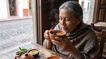 Mujer de pelo gris con chal estampado sostiene una taza de barro humeante con rodaja de cítrico y palo de canela en una mesa de madera.