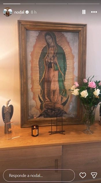 Un altar casero con una imagen enmarcada de la Virgen de Guadalupe, flanqueada por un ramo de flores y una vela encendida, sobre un mueble de madera