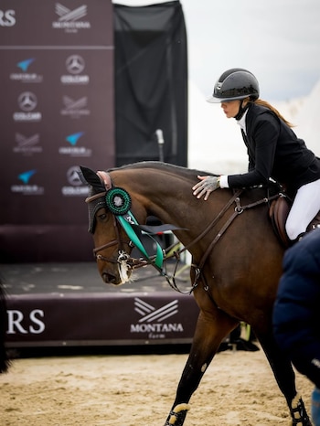 Una jinete con casco acaricia a un caballo marrón con una roseta verde en la brida, en un evento hípico con logos de patrocinadores en el fondo