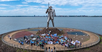 Estatua metálica de un guerrero con lanza en una plataforma circular junto a un río, con una multitud de personas y una ciudad en la distancia