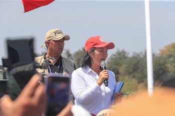 Mujer con gorra roja y camisa blanca habla por micrófono, hombre con gorra beige de policía detrás, manos con teléfonos en primer plano, bandera roja, poste blanco, árboles, cielo despejado
