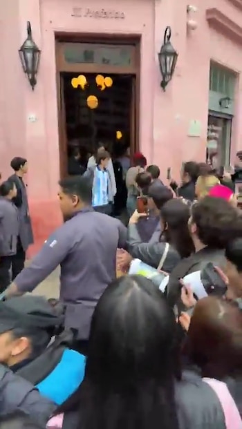 Multitud de personas y personal de seguridad frente a un edificio rosado con puertas abiertas; una persona usa camiseta de la selección argentina