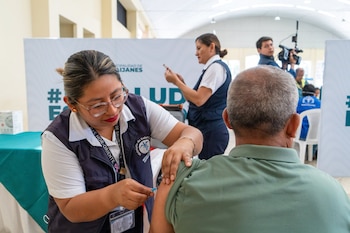 Mujer con gafas y chaleco aplica inyección en el brazo de un hombre. En segundo plano, otra mujer, un camarógrafo y carteles con texto