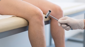 Una mano con guante blanco sostiene un martillo metálico de percusión frente a la rodilla de una persona sentada sobre una camilla.