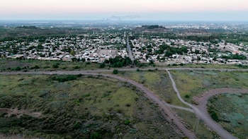 Vista aérea de un vasto terreno con vegetación baja y caminos de tierra que se extiende hacia una zona urbana densamente poblada bajo un cielo claro