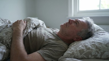 Un hombre de unos 60 años con cabello gris yace boca arriba en su cama, cubierto por sábanas estampadas. Mira hacia arriba con expresión seria.