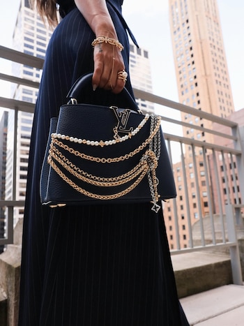 Primer plano de una mujer sujetando un bolso negro Louis Vuitton decorado con cadenas doradas y perlas, luciendo joyas y un vestido a rayas en Nueva York