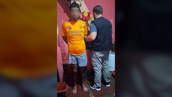 Joven con camiseta naranja y shorts frente a policía en habitación de paredes rosadas. Adorno floral en pared y cubos en suelo