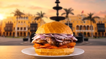 Sándwich de chicharrón con camote y cebolla en plato blanco sobre una mesa, con una plaza colonial desenfocada al fondo.