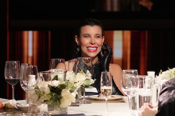 Charlotte Caniggia, con vestido y guantes negros, sonríe sentada a una mesa servida con copas, platos y arreglos florales
