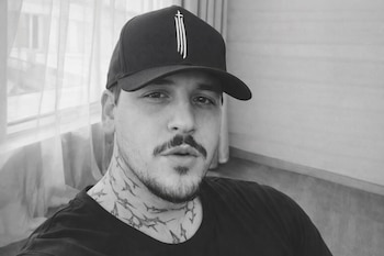 Primer plano en blanco y negro de un hombre con gorra negra, bigote, barba y tatuajes que parecen tiburones en el cuello.