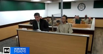 Una mujer con uniforme beige y un hombre de traje en primer plano, detrás una barandilla y otros reclusos sentados en un tribunal