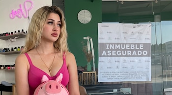 Una mujer joven rubia con top rosa posa en un salón con paredes verdes; al fondo se ve un letrero grande de "Inmueble Asegurado" en la puerta de cristal.