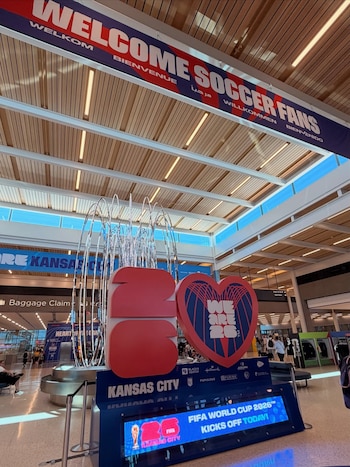 Interior de aeropuerto decorado para el Mundial 2026. Un cartel 'WELCOME SOCCER FANS', un gran '26' rojo, un corazón y una pantalla digital anunciando la FIFA World Cup 2026