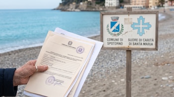 Mano sostiene papeles oficiales frente a una playa de guijarros con el mar. Un cartel muestra logos del Comune di Spotorno y Suore di Carità di Santa Maria.