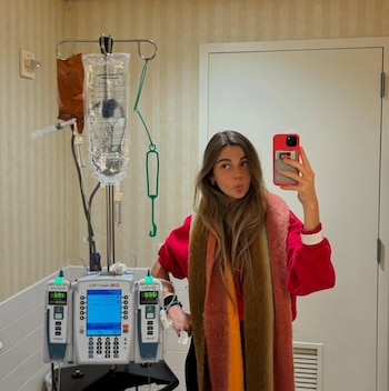 Joven mujer con top rojo y bufanda multicolor toma selfie con teléfono rojo junto a un poste con suero y un dispositivo médico de infusión