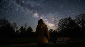 Una mujer de espaldas, con chaqueta marrón, mira hacia el cielo oscuro y estrellado donde se ve la Vía Láctea en un parque arbolado de noche.