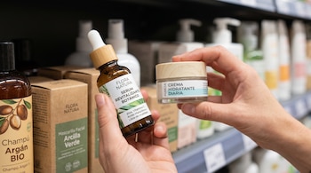 Manos sostienen un sérum revitalizante y una crema hidratante. En el fondo, estantes de supermercado con envases de productos de belleza.