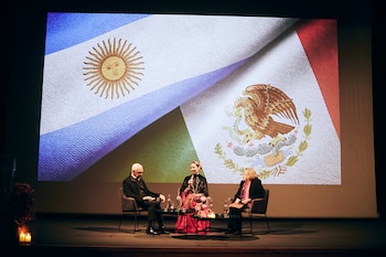 Tres personas sentadas en sillas en un escenario, una mujer habla por micrófono. Detrás, una pantalla muestra las banderas de Argentina y México. Mesa con flores, velas