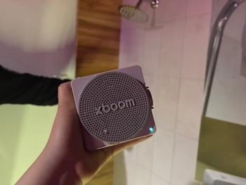Una mano sostiene un altavoz cuadrado con la inscripción xboom y gotas de agua, frente a azulejos y un cabezal de ducha