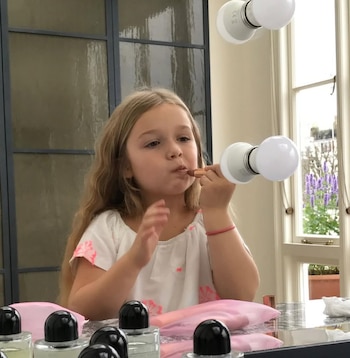 Una niña rubia con camisa blanca aplica lápiz labial. Se refleja en un espejo con luces y se ven frascos de maquillaje en la mesa