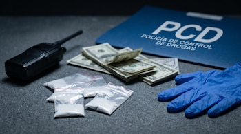 Vista detallada de una mesa con cuatro bolsas de droga, fajos de billetes, un walkie-talkie negro, guantes de látex azules y una carpeta de la PCD.