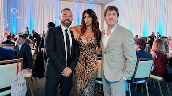 Leandro Barbuscio (esmoquin), Judith Hofman (vestido de leopardo) y Opy Morales (traje claro) posan en una gala formal. Fondo con cortinas blancas iluminadas en azul y globos