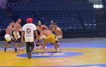 Varios hombres juegan kabaddi en una cancha amarilla y azul. Un oficial con turbante rojo camina en primer plano, y las gradas vacías del estadio se ven al fondo