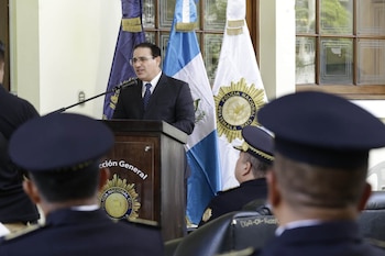 Hombre con traje y corbata habla en un podio con micrófono. Hay dos banderas de Guatemala y una bandera azul. Varios agentes de policía con uniforme y gorra observan