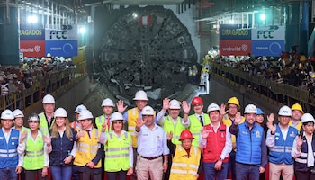 Grupo de personas con cascos y chalecos de seguridad celebrando ante la cabeza de una tuneladora que perfora un muro. Público en balcones superiores y logos de empresas