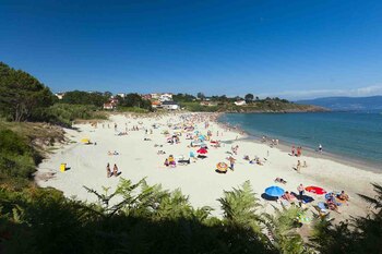 Vista panorámica de una playa de arena con muchas personas, sombrillas coloridas, mar, costa con vegetación y casas, y cielo azul despejado