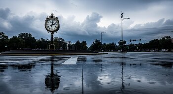 Un reloj de calle clásico con fondo de ciudad y cielo nublado. El asfalto mojado refleja las nubes. Se ven árboles, edificios y semáforos.
