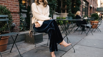Una persona sentada en una silla de metal viste un pantalón de terciopelo azul, un suéter beige y zapatos de tacón alto junto a una mesa.