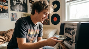 Un joven de pelo castaño claro sonriendo mientras inserta un CD en un reproductor de música plateado en su habitación con pósters y vinilos en la pared.
