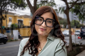 Mujer con cabello oscuro y lentes negros posa sonriendo, con un collar de corazón, en un entorno urbano con árboles y edificios de fondo