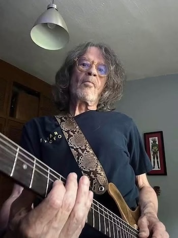 Hombre con cabello largo y gafas tocando una guitarra eléctrica con correa de serpiente; lámpara de techo y cuadro en la pared