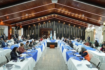 Personas sentadas a lo largo de mesas con manteles blancos y azules en un salón con techo de madera y la bandera de Guatemala al fondo