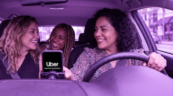 Vista interior de un coche con filtro morado, tres mujeres sonriendo: una conductora y dos pasajeras. La conductora sujeta un teléfono con el logo de Uber visible.
