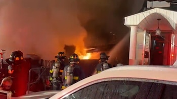 Imagen nocturna de tres bomberos con equipo completo rociando agua sobre un gran incendio con llamas y humo espeso que emerge de un edificio