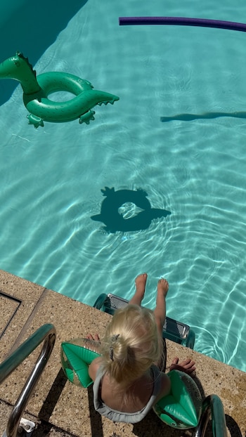 Vista aérea de niña rubia con flotadores de brazo sentada al borde de una piscina, donde flotan un dragón inflable verde y un tubo morado