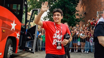 Joven con camiseta roja saluda con la mano. Una credencial está en su cuello. Está junto a la puerta abierta de un autobús rojo con estrellas. Detrás, una multitud.
